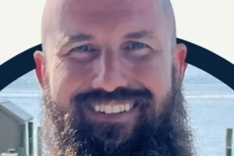 Smiling bald man with a long beard wearing a blue Columbia shirt near a waterfront dock on a clear day.