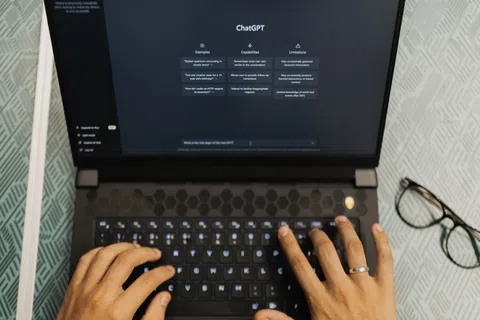 Hands typing on a laptop displaying the ChatGPT interface with examples, capabilities, and limitations sections.
