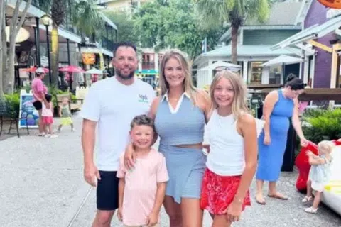 Smiling family of four posing outdoors on a sunny day in a lively shopping area with palm trees and shops.
