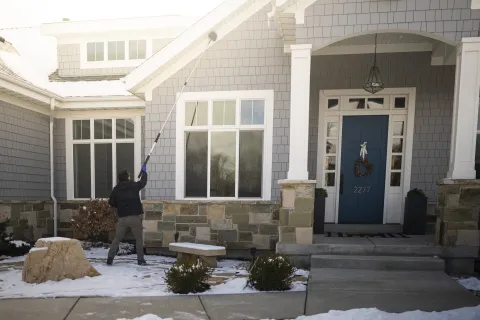 Person using a long tool to clean windows of a gray house with snow on the ground