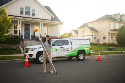 Woman with cleaning equipment standing in front of a Greenix pest control truck on a suburban street.