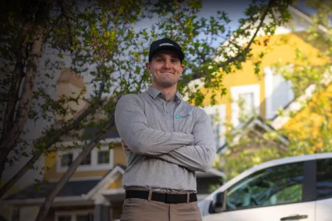 Confident technician with crossed arms standing outdoors near a white service truck and yellow house in sunlight.