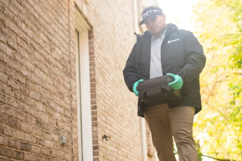 Person wearing gloves and tech gear holding rodent box walking outside home covered in leaves.