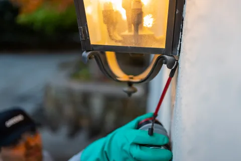 Person wearing green gloves using a screwdriver to fix an outdoor wall-mounted lantern light at dusk.