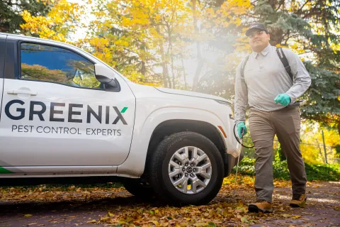 Pest control technician walking with equipment near a Greenix Pest Control Experts vehicle in autumn setting.