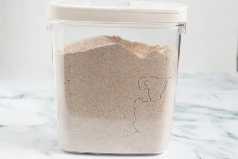 Clear plastic container filled with whole wheat flour on a marble countertop with a white lid.