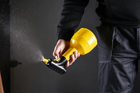 Person holding a yellow spray nozzle releasing a fine mist against a dark background.