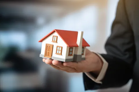 Person holding a small model house with a red roof, symbolizing real estate or home ownership.