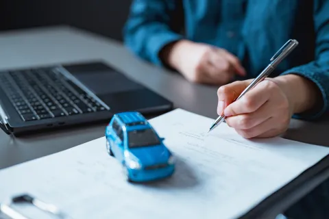 Person signing documents next to a toy blue car and a laptop, symbolizing car insurance or auto contract.
