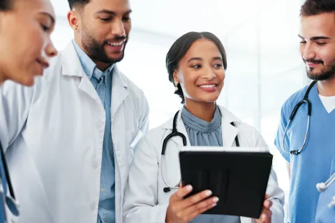 Smiling diverse medical team discussing patient data on tablet in bright hospital setting.