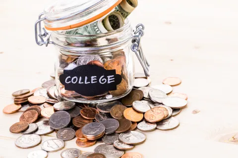 Glass jar labeled college filled with coins and rolled dollar bills on wooden surface with scattered coins