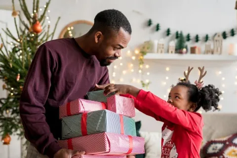 Father holding stacked wrapped Christmas gifts while his daughter reaches for presents wearing reindeer antlers