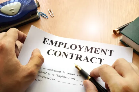 Hands holding a pen and reviewing an employment contract document on a wooden desk with office supplies.