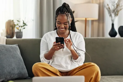 Smiling young woman sitting cross-legged on sofa using smartphone in bright living room with plants.