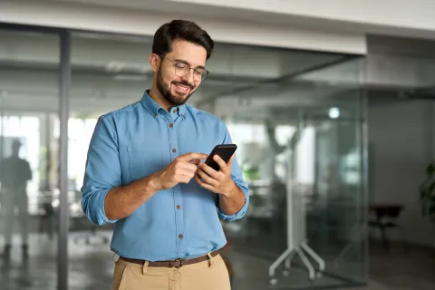 Smiling man in blue shirt using smartphone in modern office with glass walls and blurred background