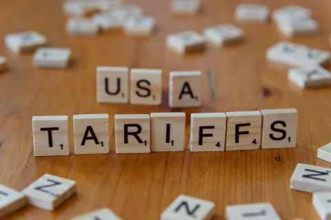 Wooden Scrabble tiles spelling USA TARIFFS on a wooden table with scattered tiles around.