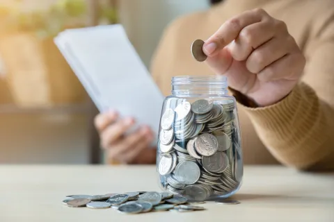 Person placing a coin into a jar full of coins while holding a notebook, symbolizing saving money.
