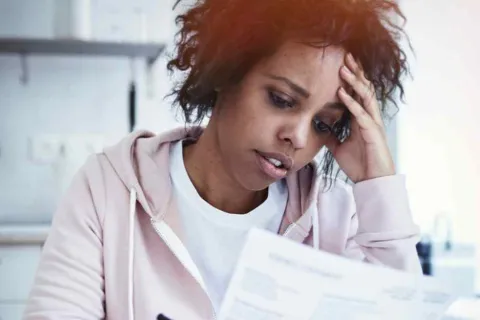 Worried woman reviewing bills at home, holding her head with a stressed expression in a bright kitchen setting