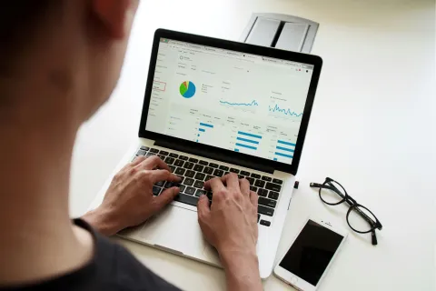 Person analyzing charts and graphs on laptop with smartphone and glasses on white desk