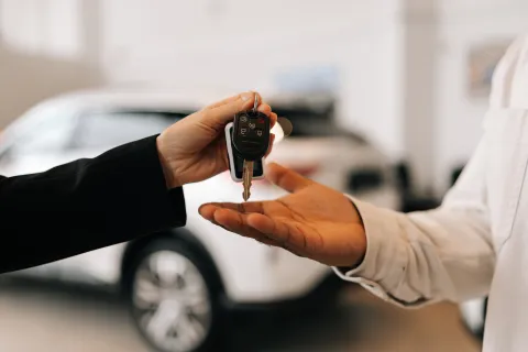 Person handing over car keys to another person with blurred white car in the background inside a showroom.