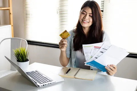 Smiling woman holding credit card and bills while working on laptop in bright home office setting.