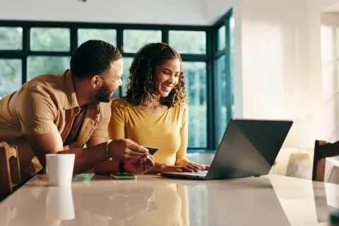 Couple shopping online with laptop and credit card in bright kitchen, smiling and enjoying the experience