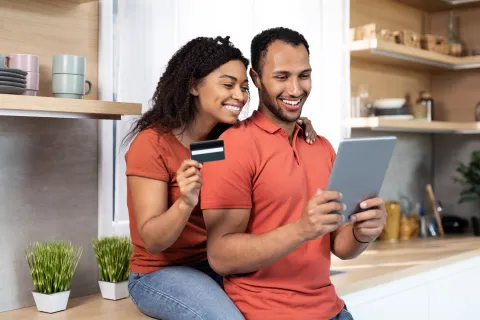 Smiling couple using a tablet and holding a credit card in a bright modern kitchen for online shopping.