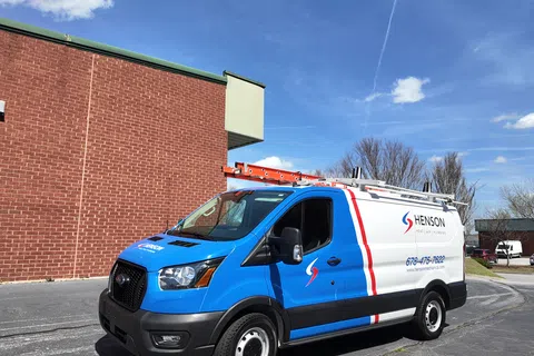 White and blue Henson service van with ladder rack parked beside a brick building under blue sky.