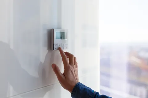 Hand adjusting modern digital thermostat on white wall with visible temperature display.
