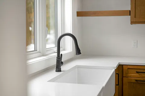 Modern kitchen sink with matte black faucet, white countertop, and wooden cabinets under natural light.