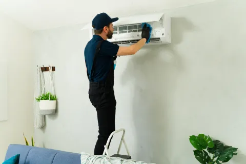 Technician in uniform servicing a wall-mounted air conditioner unit on a gray wall inside a living room.