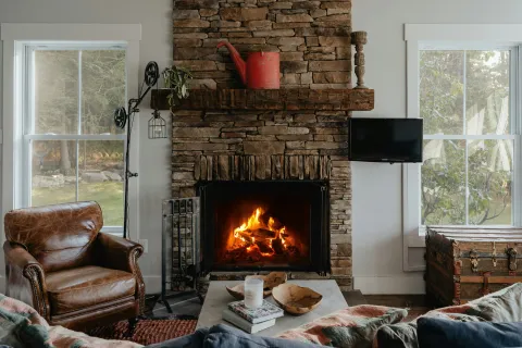 Cozy living room with stone fireplace, burning fire, leather chair, coffee table, and large windows with outdoor views