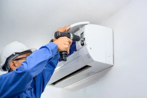 Technician in blue uniform and white helmet installing or repairing a white wall-mounted air conditioning unit with a electric drill.