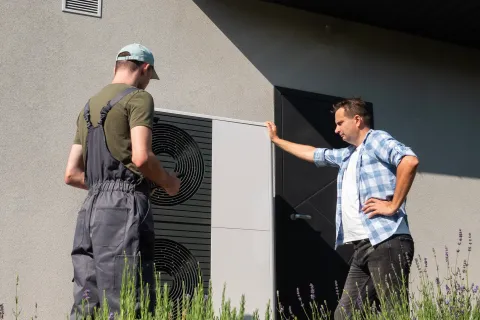 Technician and homeowner inspecting an outdoor heat pump unit next to a modern house exterior in daylight.