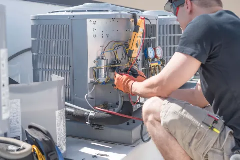 Technician wearing gloves repairing an outdoor HVAC unit using electrical tools on a sunny day.