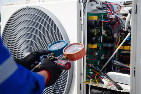 Technician in gloves using gauges to check and repair an air conditioning unit's electrical components.