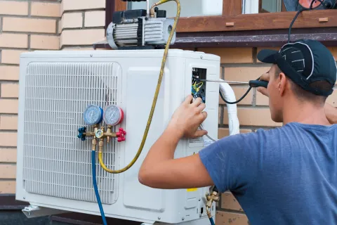 Technician servicing an outdoor air conditioning unit with gauges and tools near a brick wall.