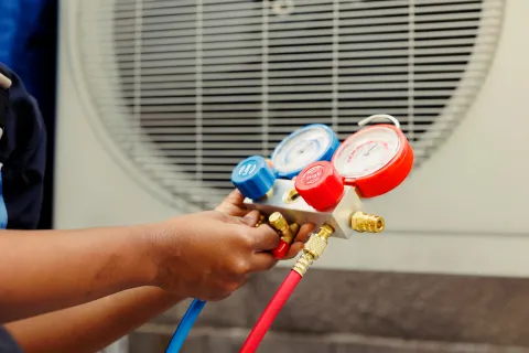 Technician's hand adjusting HVAC manifold gauge with blue and red hoses near an air conditioning unit outdoors.