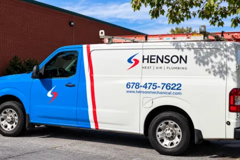 Blue and white Henson service van with logo, contact number, and ladder on top parked by a red brick building.