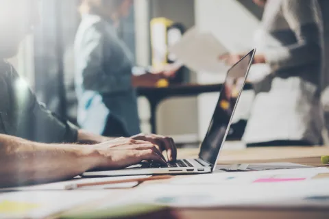 Person typing on laptop in modern office with colleagues working and documents in the background.