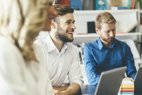 Smiling young man in white shirt during a meeting with colleagues using laptops in a modern office.