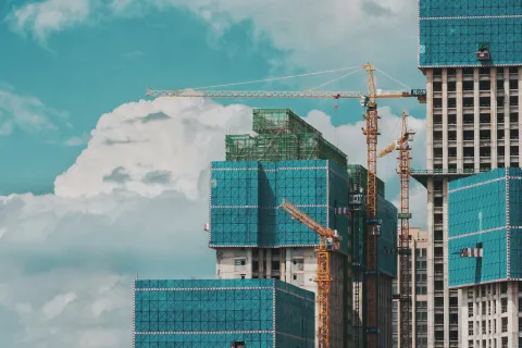 Construction cranes and modern buildings partially covered in blue safety netting against a cloudy sky.