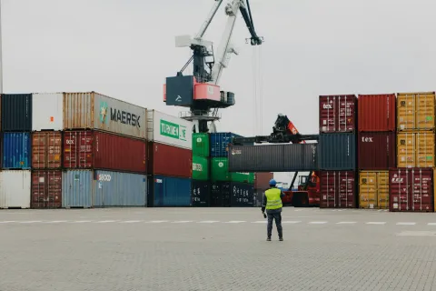 Worker in safety vest directing the loading of shipping containers at a busy industrial port with cranes.