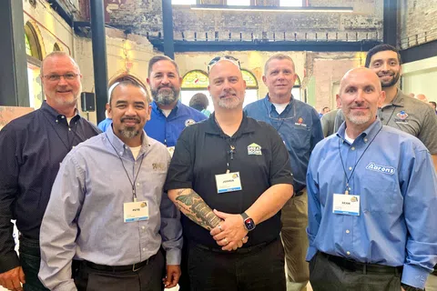 Group of seven men wearing business casual attire and name tags posing inside a rustic brick building.