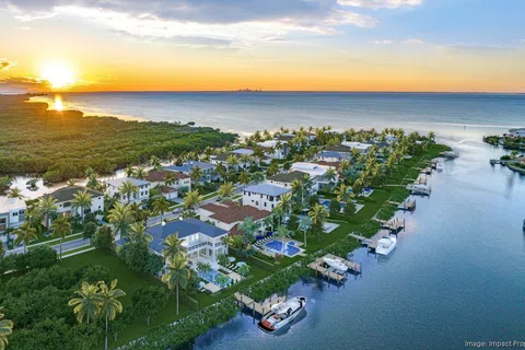 Aerial view of waterfront luxury homes with boats and palm trees at sunset along a calm waterway.