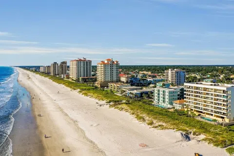 Aerial view of a beachfront city with high-rise buildings, white sandy beach, and blue ocean waves under a clear sky.