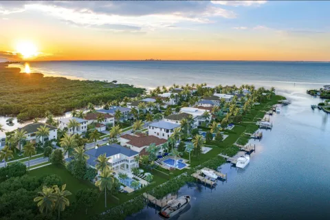 Sunset over modern waterfront homes with palm trees and boats along a canal near the ocean coastline.