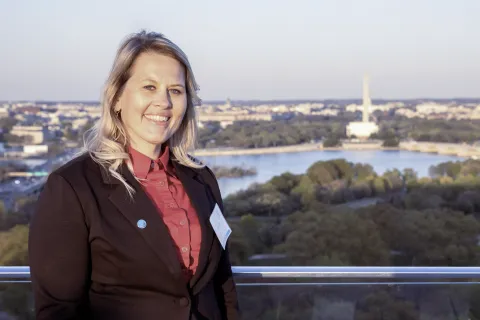 Smiling woman in business attire standing on balcony with city skyline, river, and monument in background during daylight