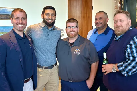 Group of five men smiling and posing indoors, dressed in business casual attire with one holding a beer bottle