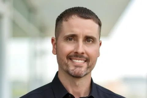 Smiling man with short hair and beard wearing a black button-up shirt with arms crossed outdoors.
